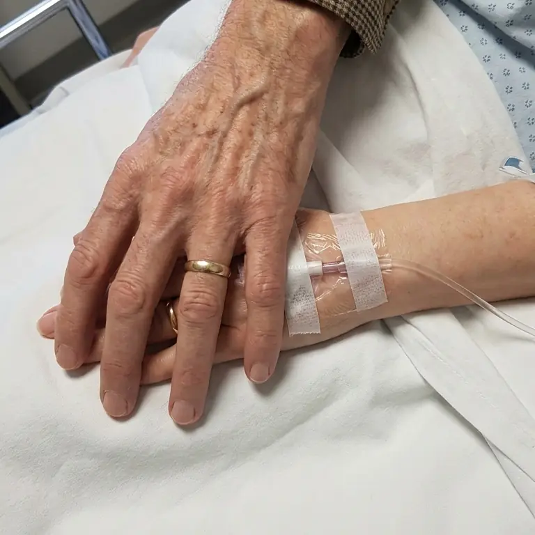 A mão de Chico, com aliança, pousada sobre a mão de Rosa, que tem um acesso venoso fixado no braço, em um leito de hospital.