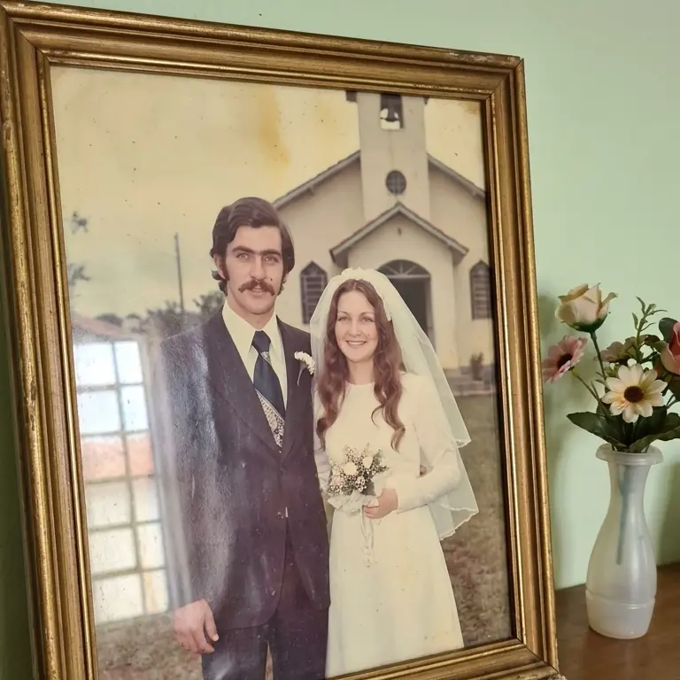 Porta-retrato dourado com uma fotografia antiga de casamento: noivo de terno escuro e noiva de véu e buquê diante de uma igreja; ao lado, um vaso com flores.